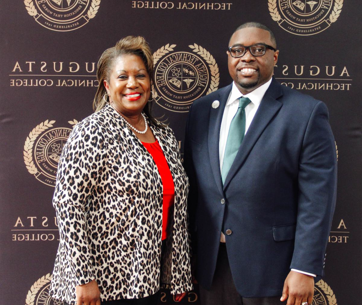 Photo: Smiling African American male wearing black glasses, 蓝色的套件, 浅蓝色/青色领带, 白领衬衫, with silver lapel pin standing next to a smiling African American female wearing red shirt, 珍珠项链, 豹纹开衫, posing for a photo in front of a black background with the 奥古斯塔 Tech seal.