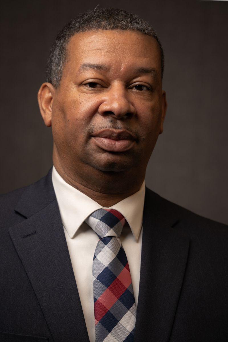 Dr. Howie Gunby, 一名非裔美国男子，身穿灰色西装，内搭白领衬衫，系一条灰色和白色相间的红色条纹格子领带.