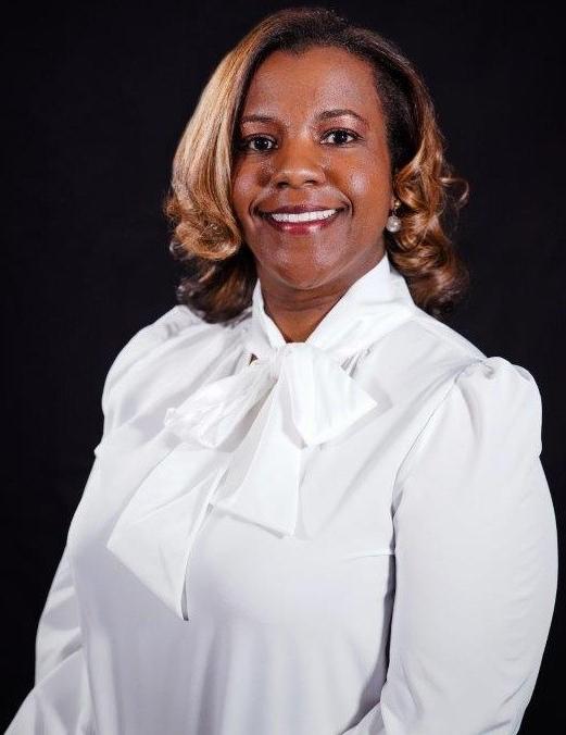 一位非裔美国女性. 克里斯蒂瑟西, 健康科学学院院长, 有一头中等长度的金色卷发，穿着白色的蝴蝶结领衬衫，戴着黑色背景的珍珠耳环，微笑着.